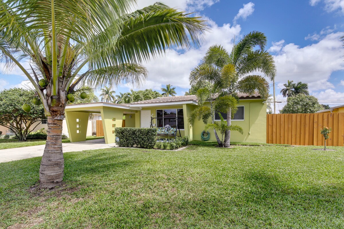 A vibrant yellow home is surrounded by lush greenery, featuring well-maintained landscaping. Palm and other trees provide shade and a tropical feel. The property is enclosed by a wooden fence, emphasizing privacy, while a spacious driveway leads to the entrance.