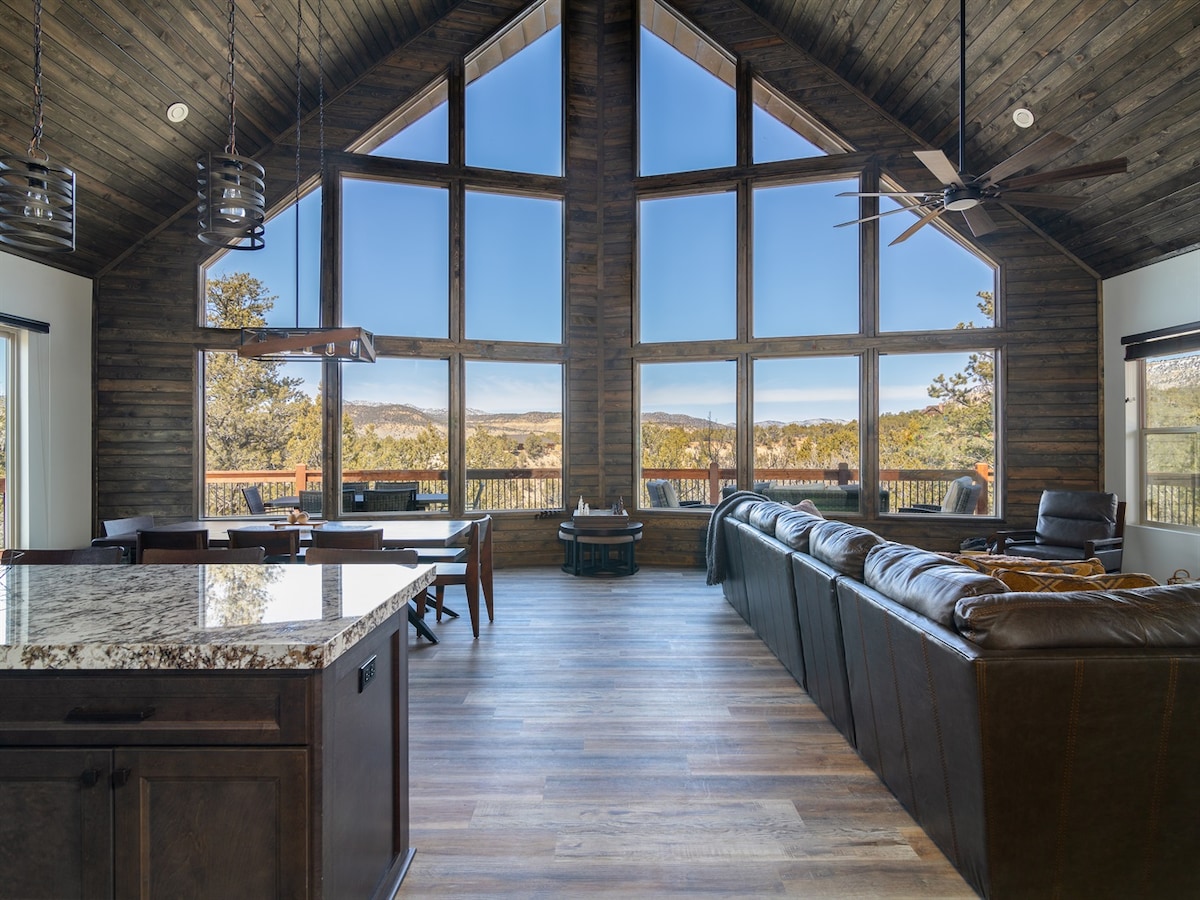 The spacious living area features vaulted wood ceilings and large windows that provide sweeping views of the surrounding landscape. A comfortable sectional sofa is situated by the windows, and a dining area is visible in the background, with a wooden table ready for gatherings.