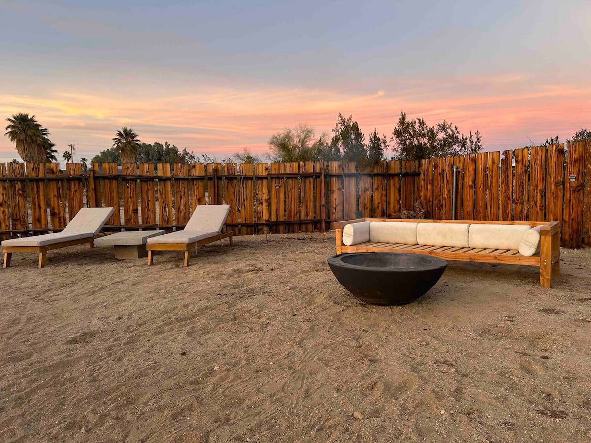 An outdoor space features a cozy seating area next to a fire pit, surrounded by a wooden fence and soft desert sand. Two sun loungers are positioned nearby, creating a relaxed atmosphere for enjoying the scenic views at sunset.