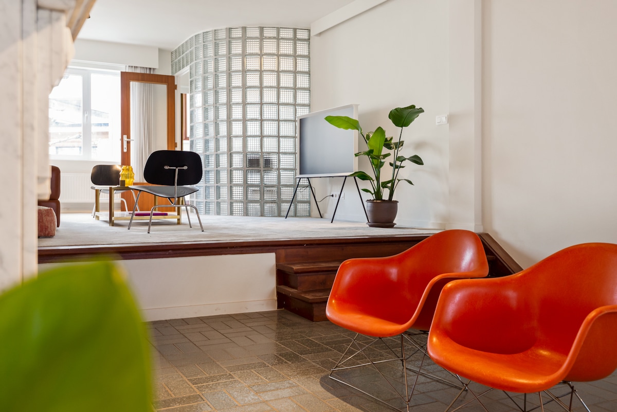 A spacious living area features striking orange chairs and a sleek coffee table. A large television is mounted on a white wall, with a glass block partition adding texture. Lush greenery is present in a potted plant, complementing the overall aesthetic.