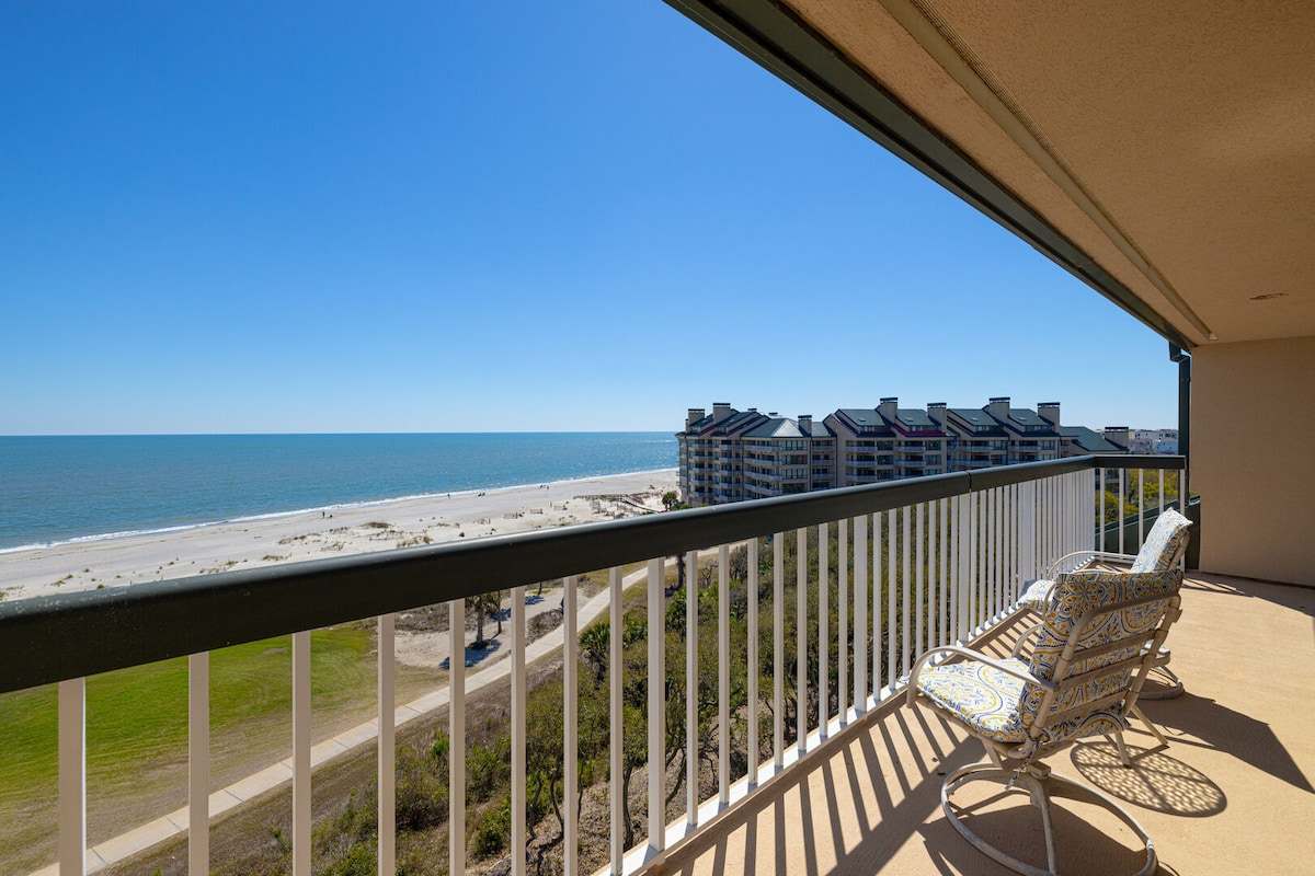 An oceanfront balcony offers sweeping views of the Atlantic Ocean and beach. A wicker chair is placed on the balcony, surrounded by a clear blue sky. The coastline is visible in the distance, framed by the resort buildings.