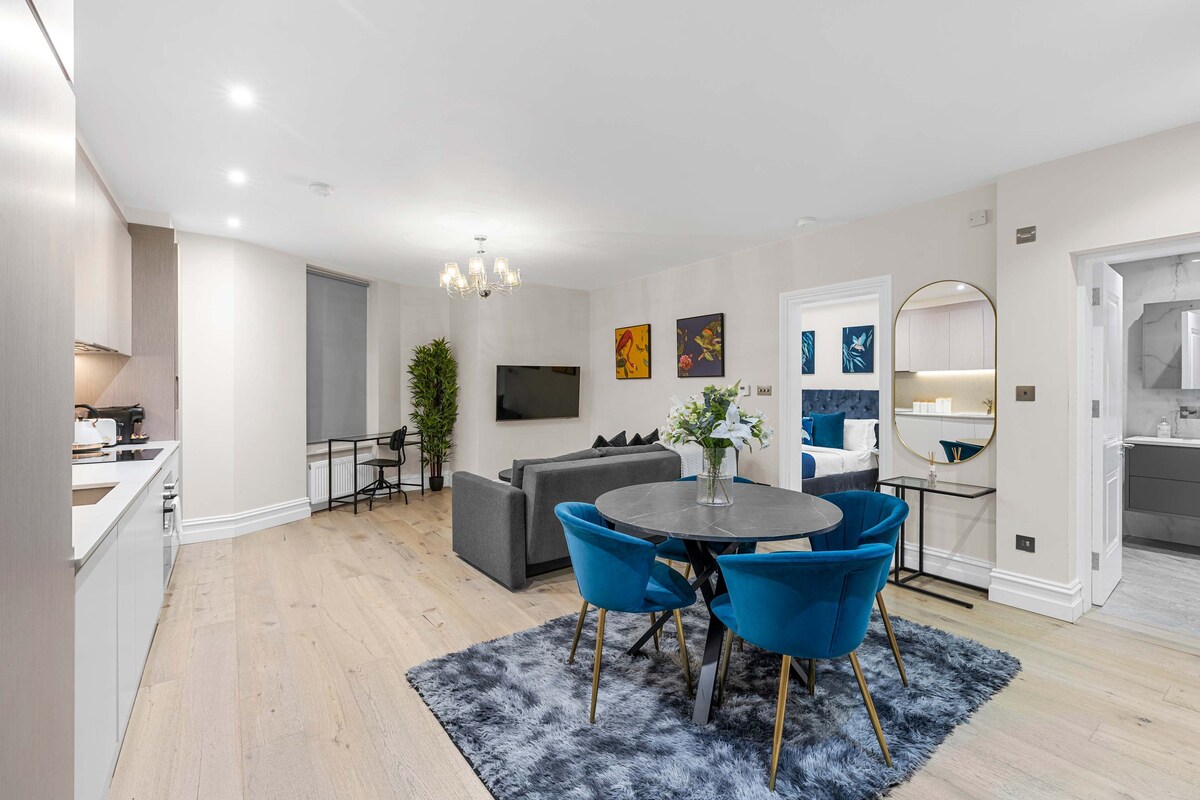 A modern living space features a round dining table surrounded by blue velvet chairs, set against a plush rug. The room has soft lighting and a large flat-screen TV mounted on the wall. Tasteful artwork adorns the walls, contributing to a stylish atmosphere.