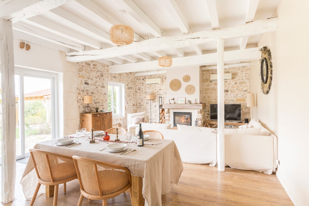 A spacious living area features natural stone walls and wooden beams, creating an airy atmosphere. A dining table is set for several guests, while a comfortable seating arrangement includes a light-colored sofa. A fireplace adds warmth, and large windows allow for abundant natural light.