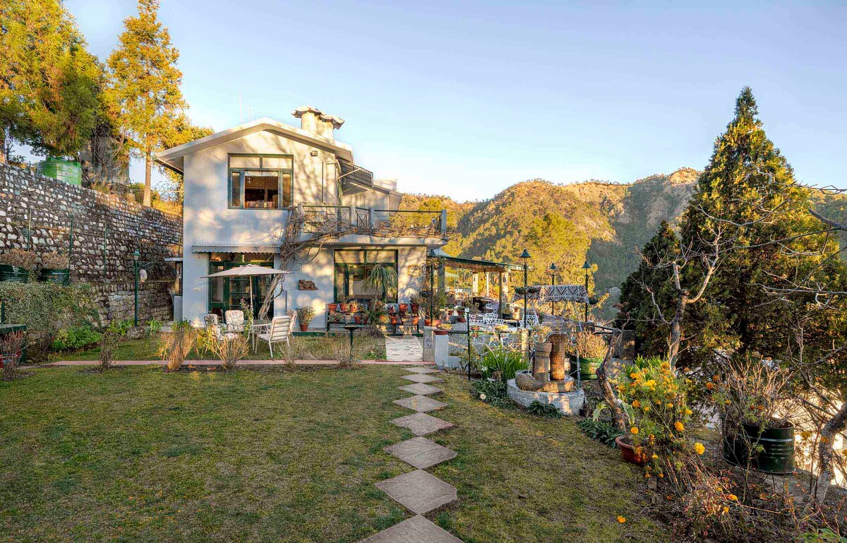 A cozy home is viewed from the lawn, showcasing multiple outdoor seating areas and large windows that invite natural light. Scenic hills create a serene backdrop, complemented by a stone pathway leading to various garden features and plant arrangements.