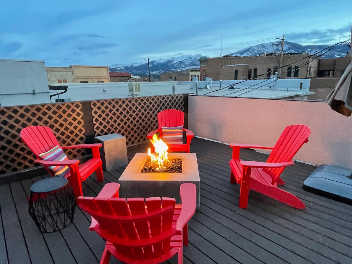 An outdoor patio features a central fire pit surrounded by four bright red Adirondack chairs and a small side table, creating a cozy gathering space. Distant mountains are visible in the background, with twilight sky casting a soft glow.