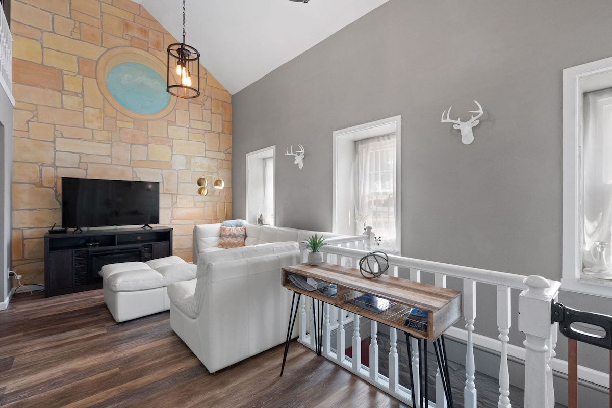 A spacious living area is highlighted by a vaulted ceiling and a stone wall with a circular mural. A comfortable white sectional sofa is positioned in front of a media console with a television. Two windows bring natural light into the room, with tasteful decor adorning the walls.