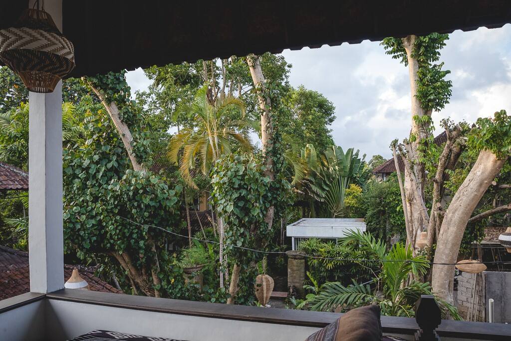 A private balcony offers lush garden views framed by tall trees and greenery. The serene setting includes a glimpse of nearby rooftops, providing a sense of tranquility amidst nature. Soft lighting highlights comfortable seating arrangements, inviting moments of reflection.