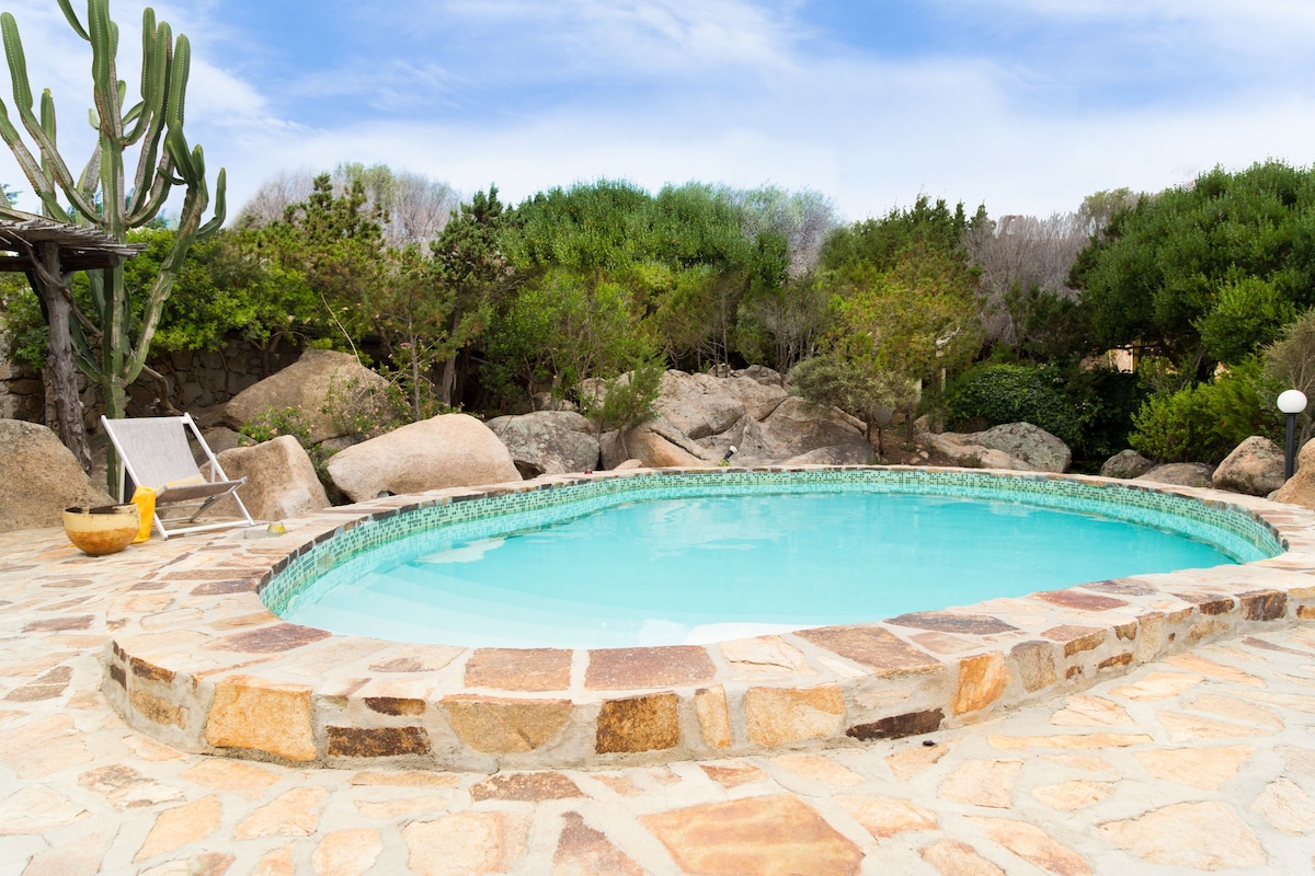 A circular swimming pool is surrounded by a stone patio, featuring natural rock formations and lush greenery. Lounge chairs are placed nearby, offering a comfortable spot to relax. The serene landscape evokes a tranquil atmosphere, inviting guests to enjoy the outdoor space.