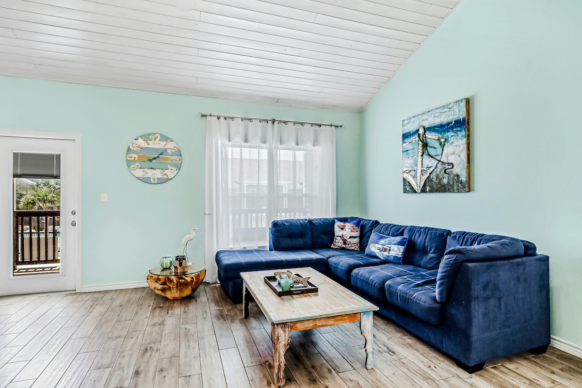 A spacious living area features soft blue walls and a comfortable sectional sofa. A rustic wooden coffee table rests in front, while light filtering through sheer white curtains enhances the relaxed atmosphere. A piece of artwork depicting a surfer adorns the wall.