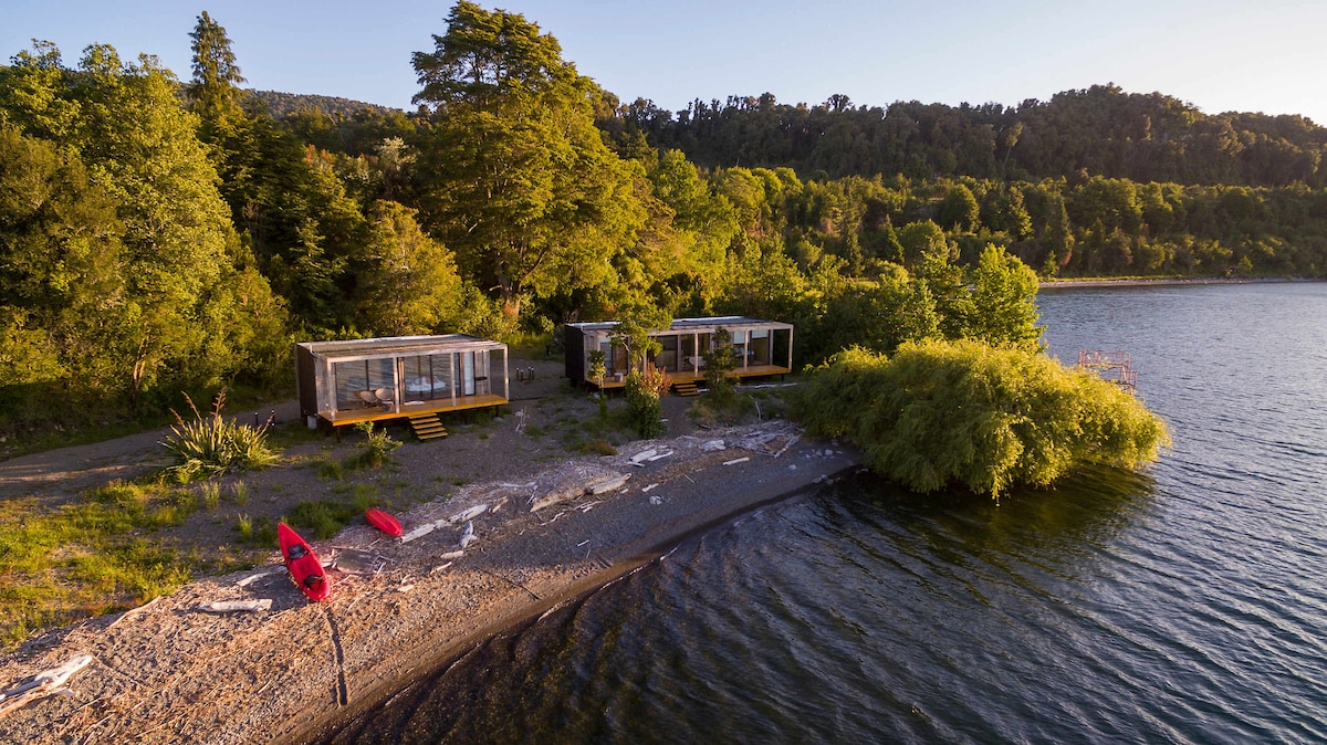 Two modern cabins are positioned along a pebbled beach, gently sloping towards a calm lake. Lush greenery surrounds the area, with a sandy stretch at the water's edge. A few kayaks rest nearby, ready for exploration of the serene landscape.