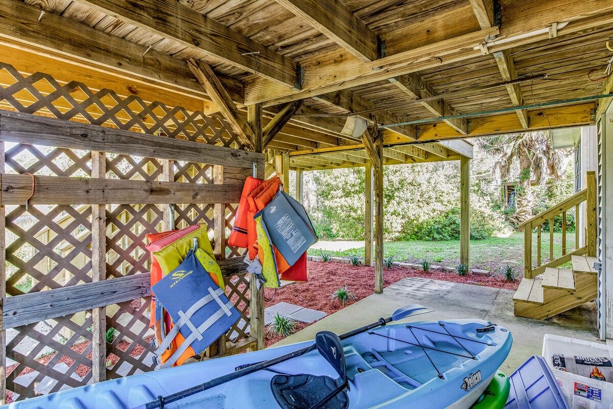 A covered outdoor area is visible, featuring two kayaks resting on the ground. Life jackets in various colors are hung on a wooden lattice. The surrounding space includes a path leading to a grassy section with landscaping.