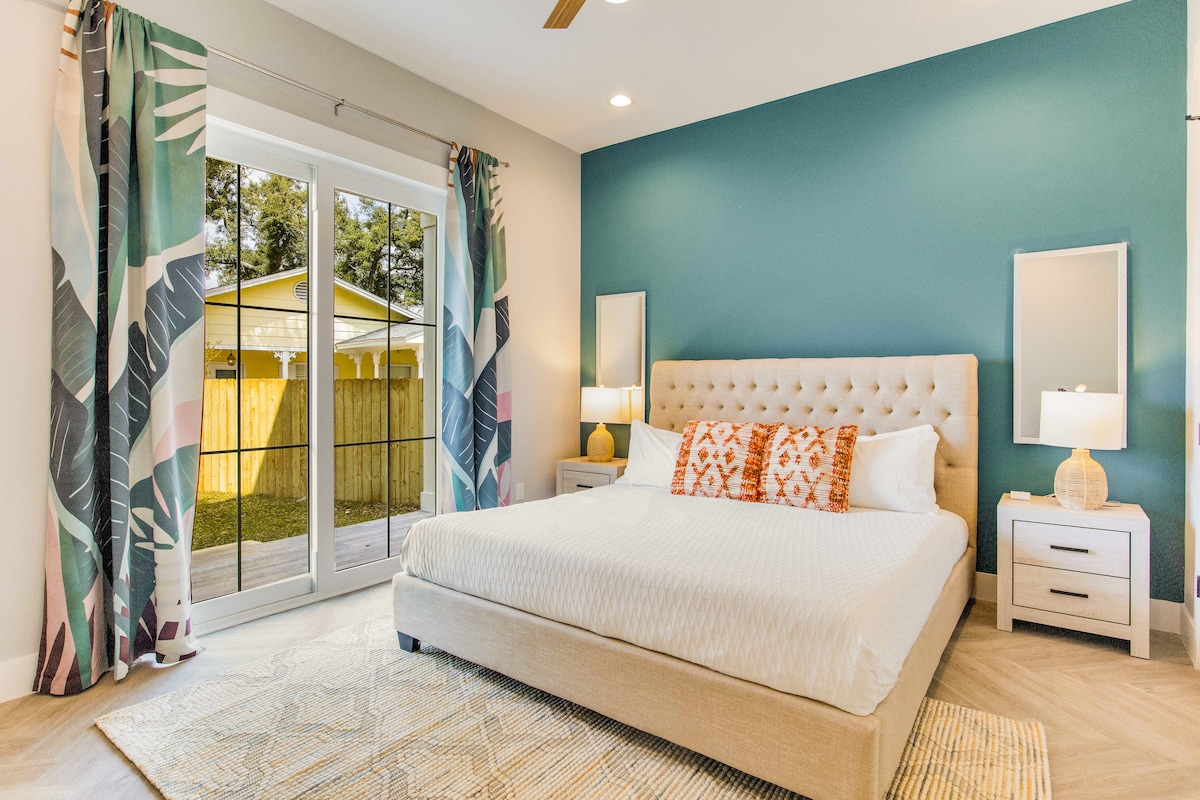 A light and airy bedroom features a spacious bed with soft linens and decorative pillows. Two bedside tables hold modern lamps, and large sliding glass doors provide natural light and a view of the outdoor space. Tropical-themed curtains frame the windows.