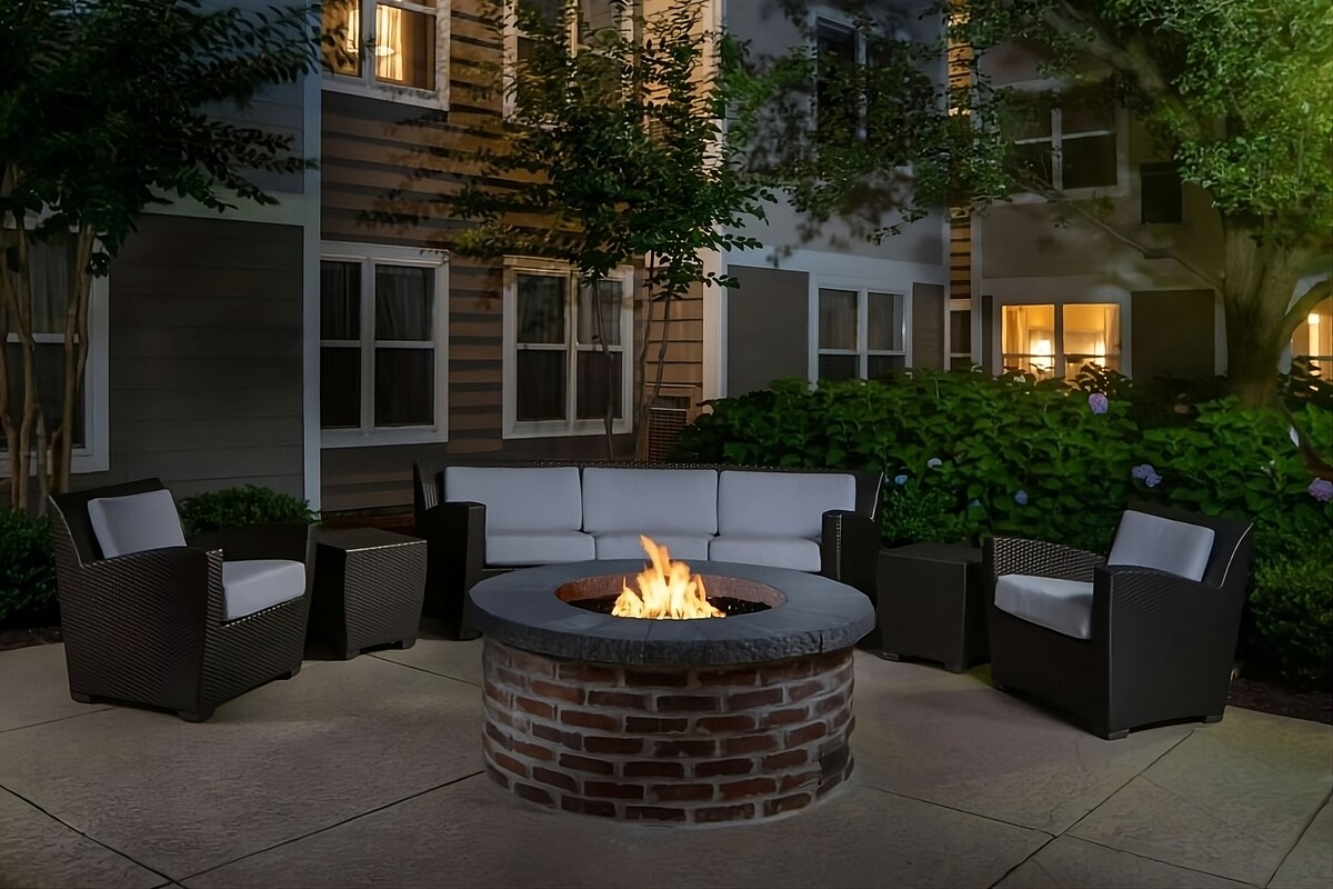An outdoor gathering area features a round stone fire pit surrounded by four comfortable lounge chairs upholstered in light fabric. Lush greenery and well-lit windows in the background create a peaceful setting for relaxation and socializing.