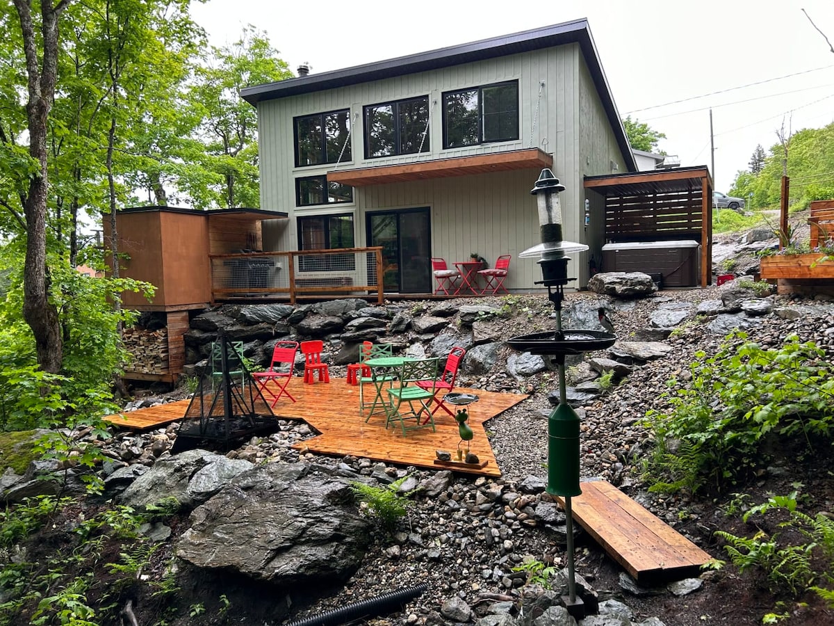 The exterior of the chalet is showcased, featuring a modern design with large windows. A wooden deck is visible, surrounded by rocky terrain, with outdoor seating in bright colors. A lantern stands nearby, contributing to the inviting outdoor space.