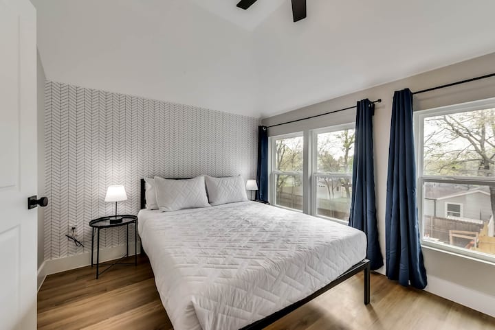  Брачен кревет (широк 150-179 см) со бела постелнина наспроти шарен ѕид. Лесниот дрвен под, ноќната масичка со ламба и големите прозорци со темносини завеси го комплетираат модерниот изглед.