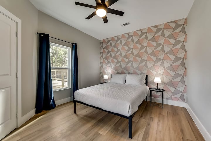 Дупли кревет со бела постелнина сместен наспроти геометриски акцентски ѕид. Опкружено со ноќни масички и светилки, со сини завеси и тавански вентилатор кој го комплетира изгледот.