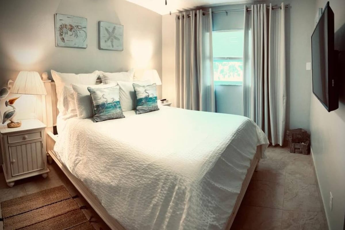 A cozy bedroom showcases a spacious bed with crisp white linens and decorative pillows. Soft lighting emanates from a bedside lamp, while light grey curtains frame a window that allows natural light to fill the room. Artwork reflecting coastal themes is displayed on the walls.