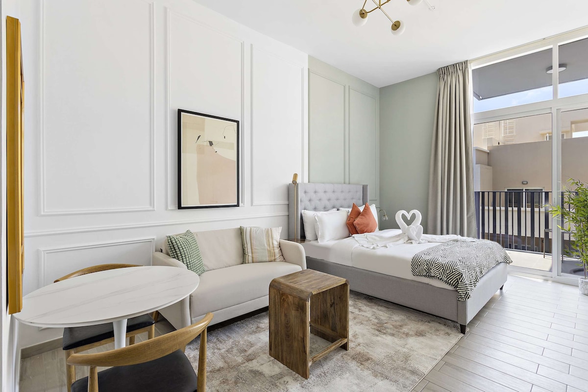 A modern studio apartment is displayed, featuring a comfortable double-sized bed with soft linens. A light-coloured sofa sits next to a charming wooden table. Large windows allow natural light to fill the space. Decorative art and a balcony view enhance the ambiance.