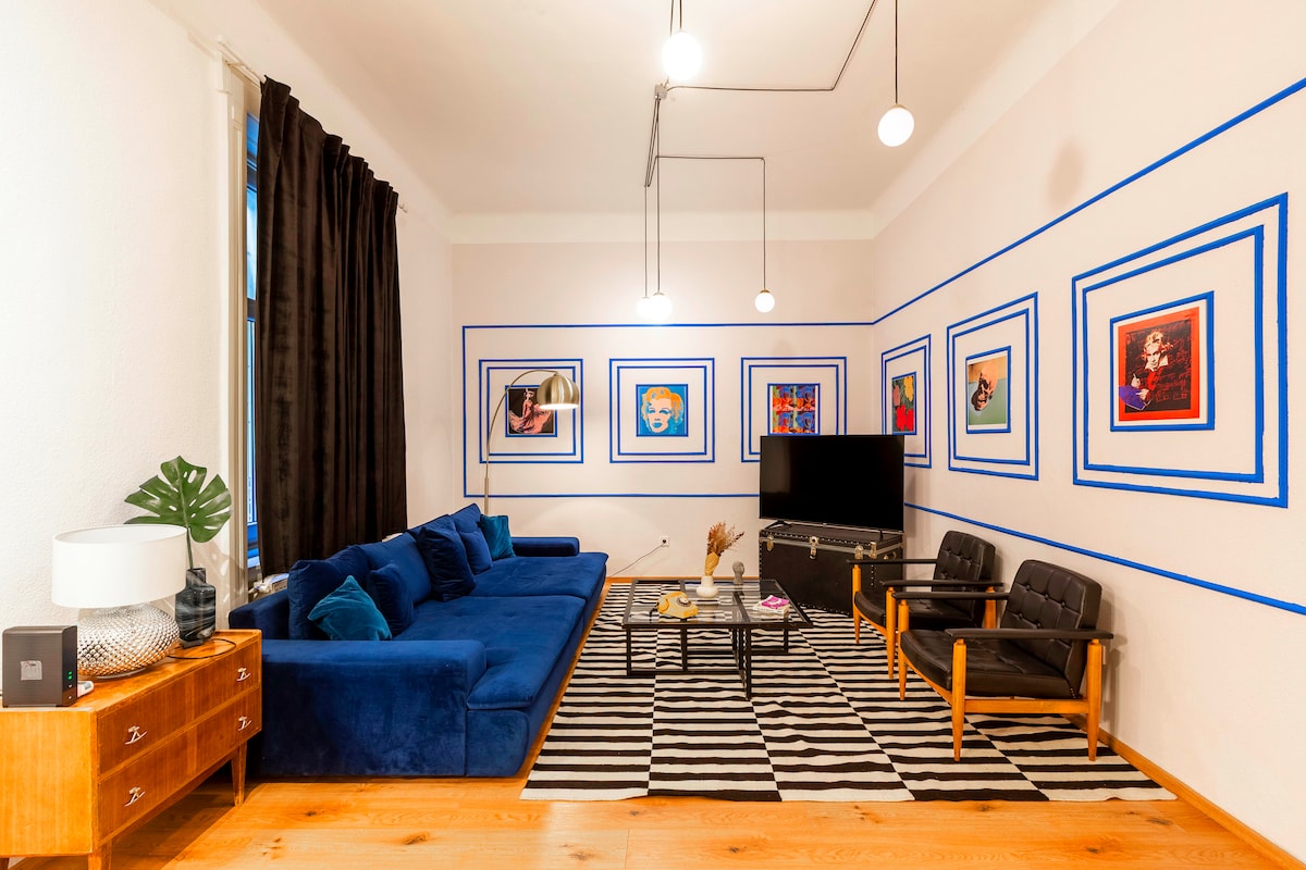 A spacious living room is highlighted by a deep blue sofa and wooden armchairs. Colorful artwork adorns the walls, framed by blue lines. A coffee table sits on a black and white striped rug, with a television positioned in front of the seating area.