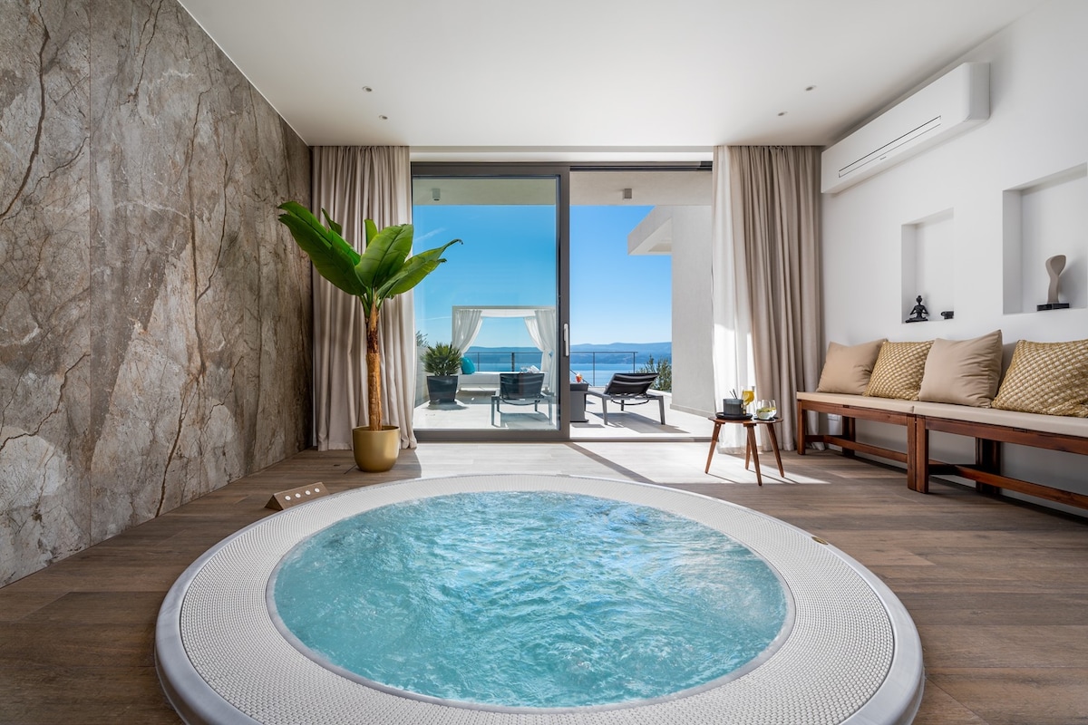 A serene spa area showcases a round hot tub with gentle water jets, inviting relaxation. Large sliding glass doors open to a sunlit outdoor terrace featuring sea views. A decorative plant adds a touch of nature, complementing the spacious and modern design of the room.