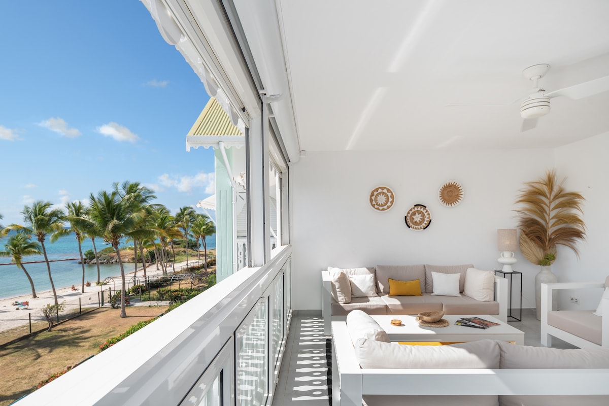 A bright sitting area is presented with a comfortable sofa, decorative wall hangings, and a serene view of palm trees and the beach. Large glass doors allow natural light to flood the space, creating an inviting connection with the outdoor scenery.