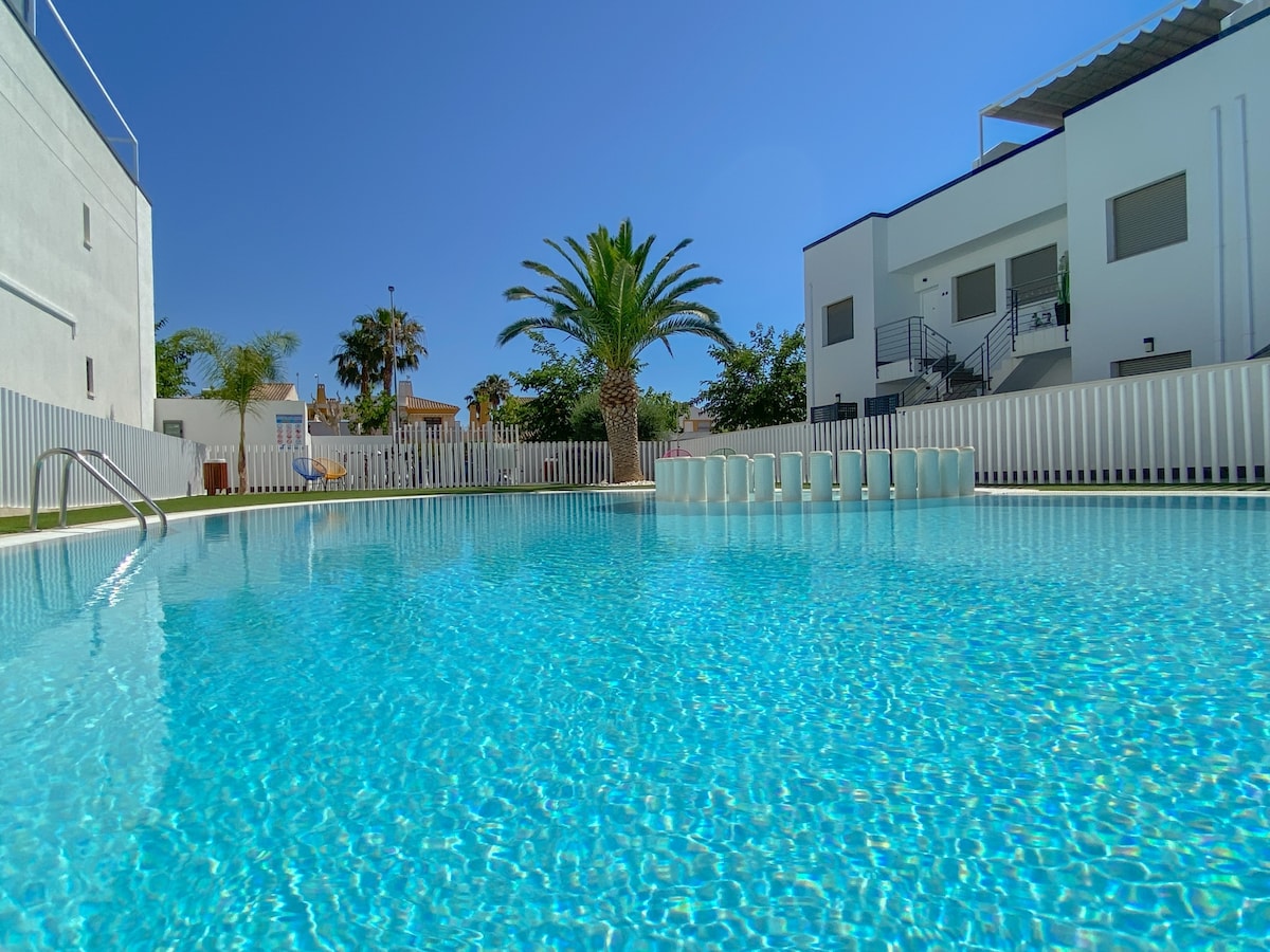 A crystal-clear swimming pool is surrounded by a sunlit area, featuring lush palm trees and outdoor seating. The pool's inviting turquoise water reflects the blue sky, while a modern white building stands in the background, providing a serene environment for relaxation.