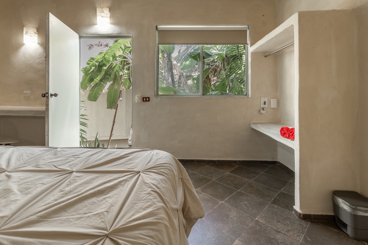 A minimalist bedroom features light-colored walls and a spacious bed with a textured comforter. Natural light floods the space through a large window, revealing tropical plants outside. A small workspace and a red towel add practical yet aesthetic touches to the room.