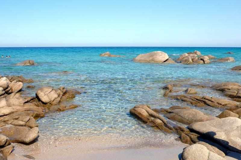A serene coastal view is presented, featuring clear turquoise water lapping against smooth, rocky formations. In the distance, gentle waves can be seen breaking on the shoreline, under a clear blue sky. The scene invites a sense of tranquility and natural beauty.