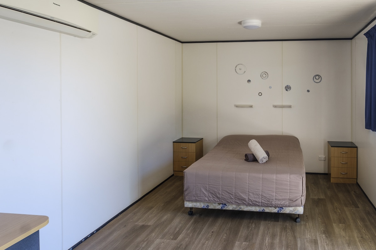 A spacious room features a large double bed with neutral bedding. Two bedside tables are positioned beside the bed, while a desk is visible in the corner. The light wooden flooring complements the minimalist design, and a window with blue curtains allows natural light to filter in.