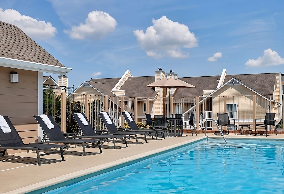 A seasonal swimming pool is surrounded by lounge chairs, with umbrellas providing shade. The area is enclosed with a fence, featuring additional seating options nearby. Light reflects off the water under a clear blue sky, while the building provides a backdrop with modern architecture.