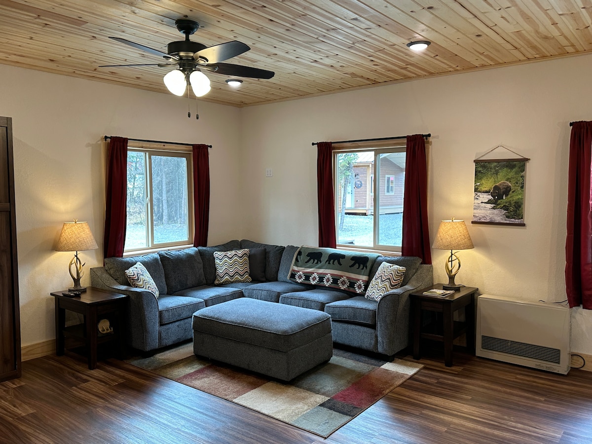 A comfortable living room features a sectional sofa accompanied by two side tables, each with a lamp. Natural light enters through two windows adorned with red curtains. A decorative area rug adds color to the hardwood floor, while a nature-themed artwork hangs on the wall.