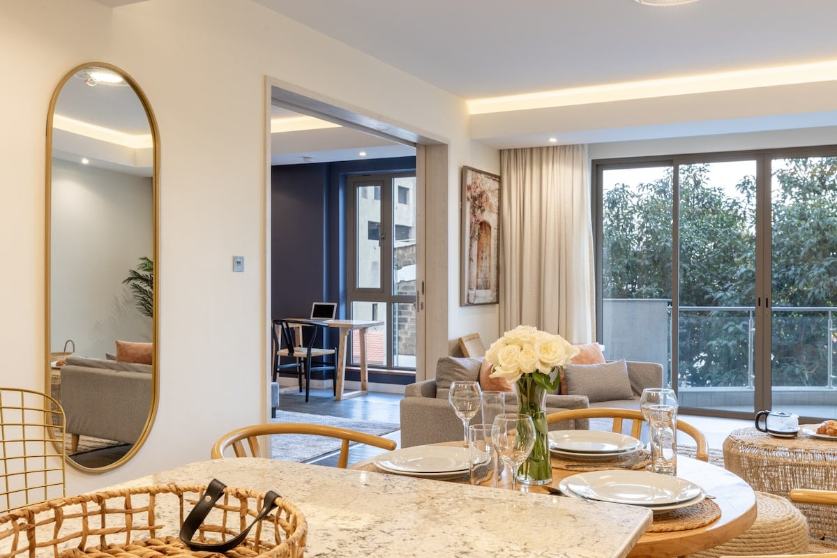 A modern living area is highlighted with soft neutral tones, featuring a round dining table with elegantly set dinnerware. A large mirror reflects natural light, enhancing the space. A comfortable sofa and armchair are positioned near sliding glass doors that open onto a balcony with views of surrounding greenery.