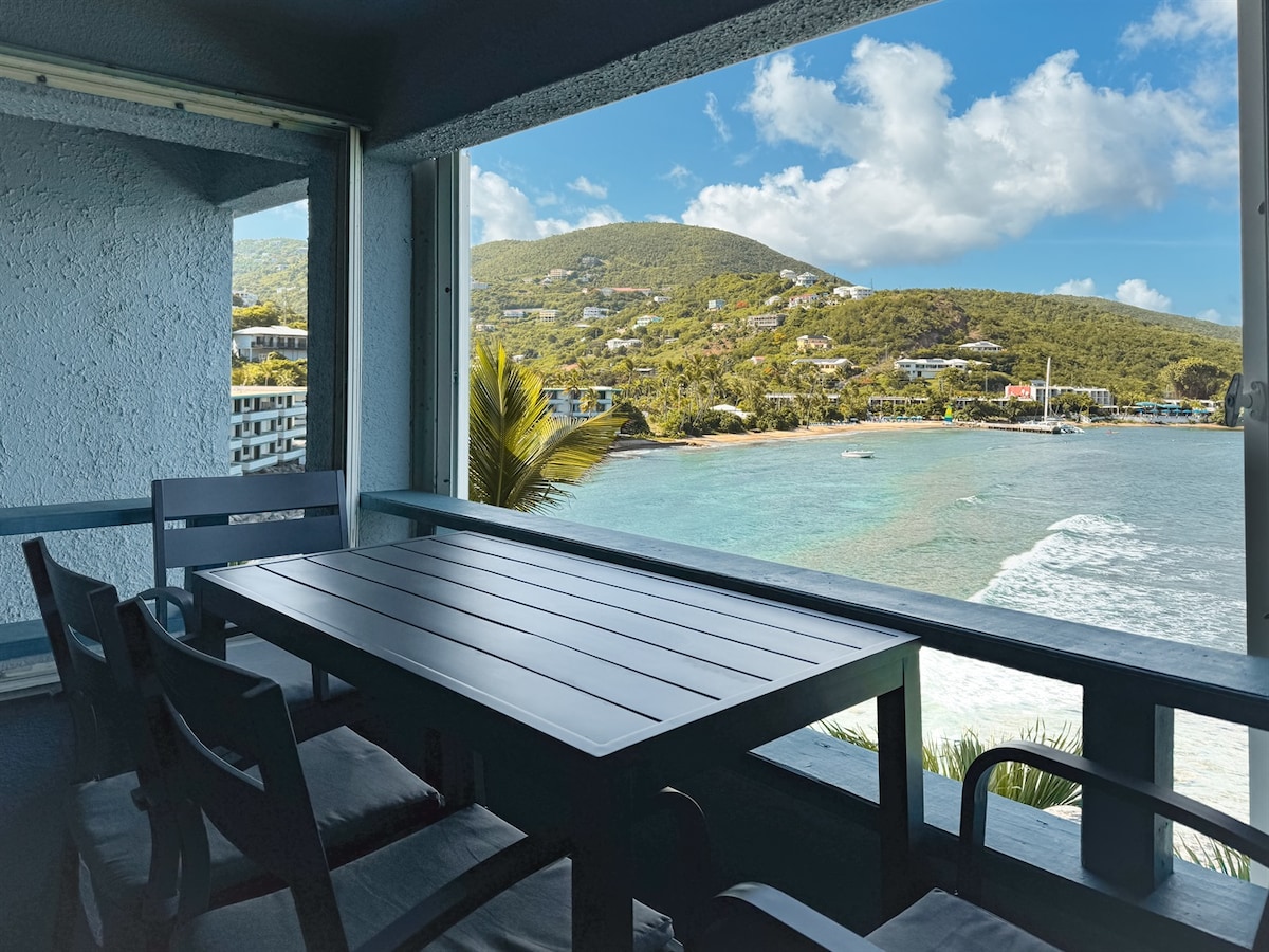 A spacious balcony features a modern dining table surrounded by four chairs, with a stunning 180-degree view of the ocean and hills in the background. Lush palm leaves frame the serene coastline, while waves gently lap against the shore.