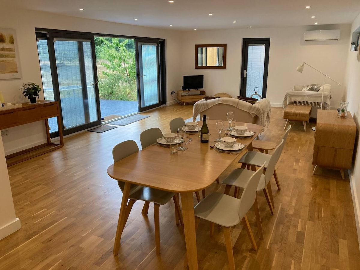 An inviting open-plan living area is featured, showcasing a wooden dining table surrounded by six chairs. A comfortable sitting area includes a television and a sofa. Large glass doors lead to the outside, allowing natural light to brighten the space.