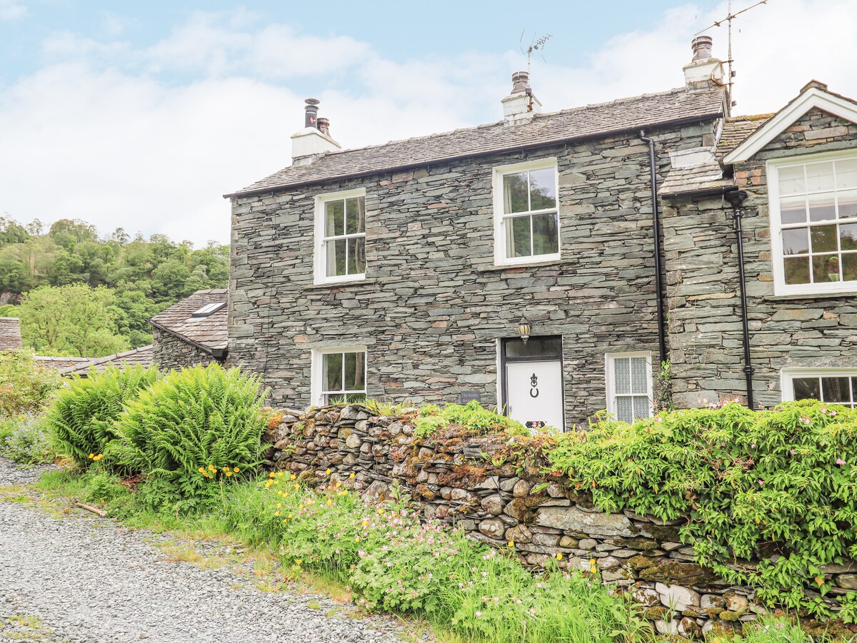 The exterior of a stone cottage features a blend of natural green foliage and well-maintained gardens. The two-story structure presents symmetrical windows and a welcoming front door, all set against a backdrop of rolling hills and lush greenery.