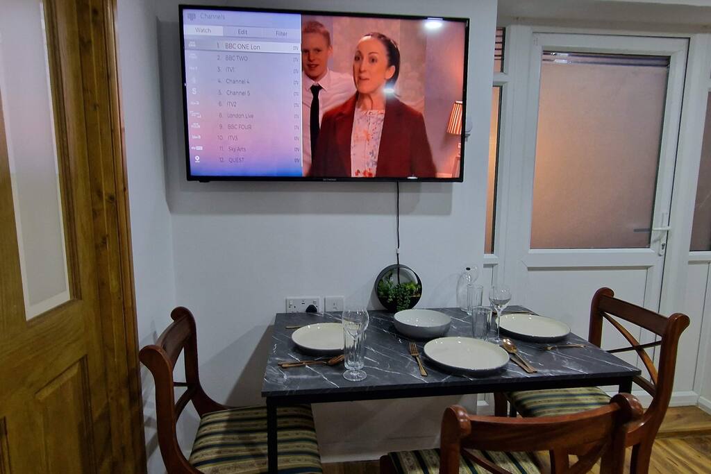 A dining area features a dark marble table set with plates, glasses, and cutlery. Behind the table, a wall-mounted television displays a program. Windows allow natural light to filter in, complemented by a potted plant on the table.