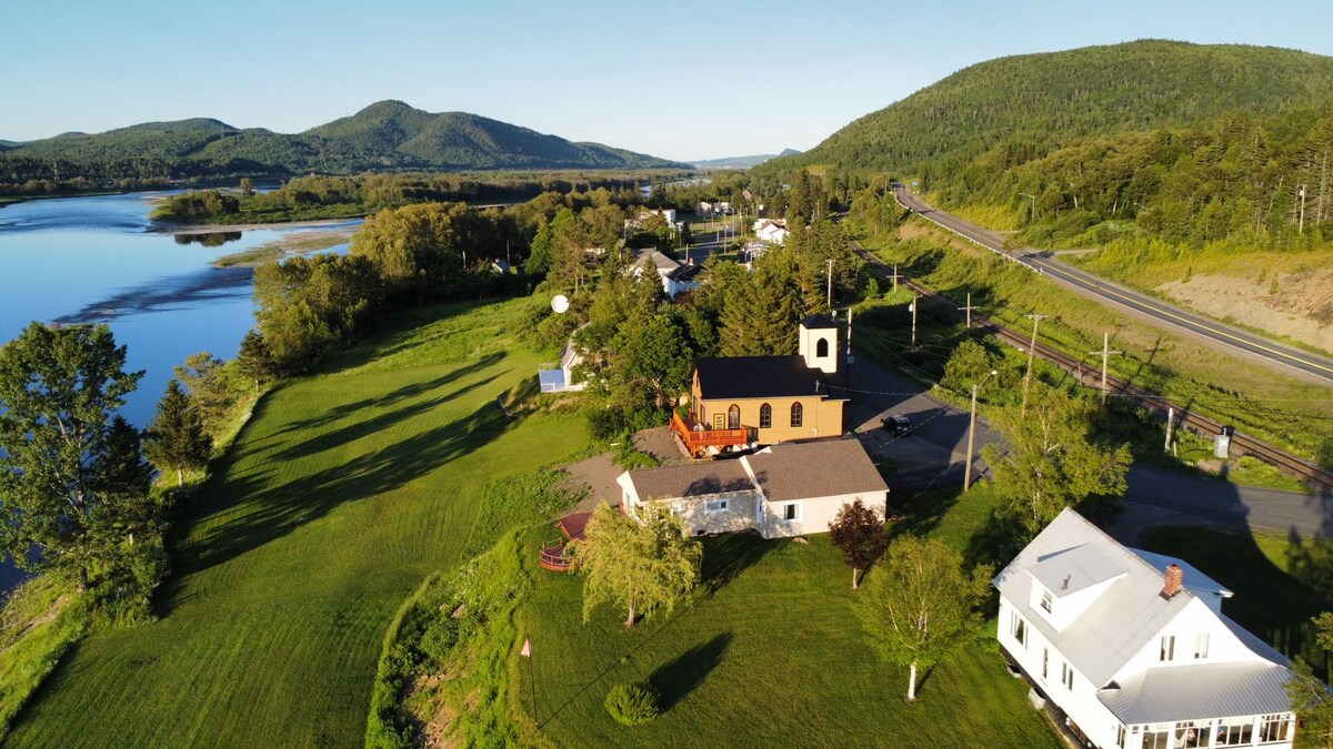 An aerial view showcases lush green landscapes surrounding the cottage, with expansive lawns and trees. The Restigouche River glimmers in the sunlight, while nearby mountains provide a scenic backdrop. The cottage is prominently situated among a few other buildings along a quiet road.