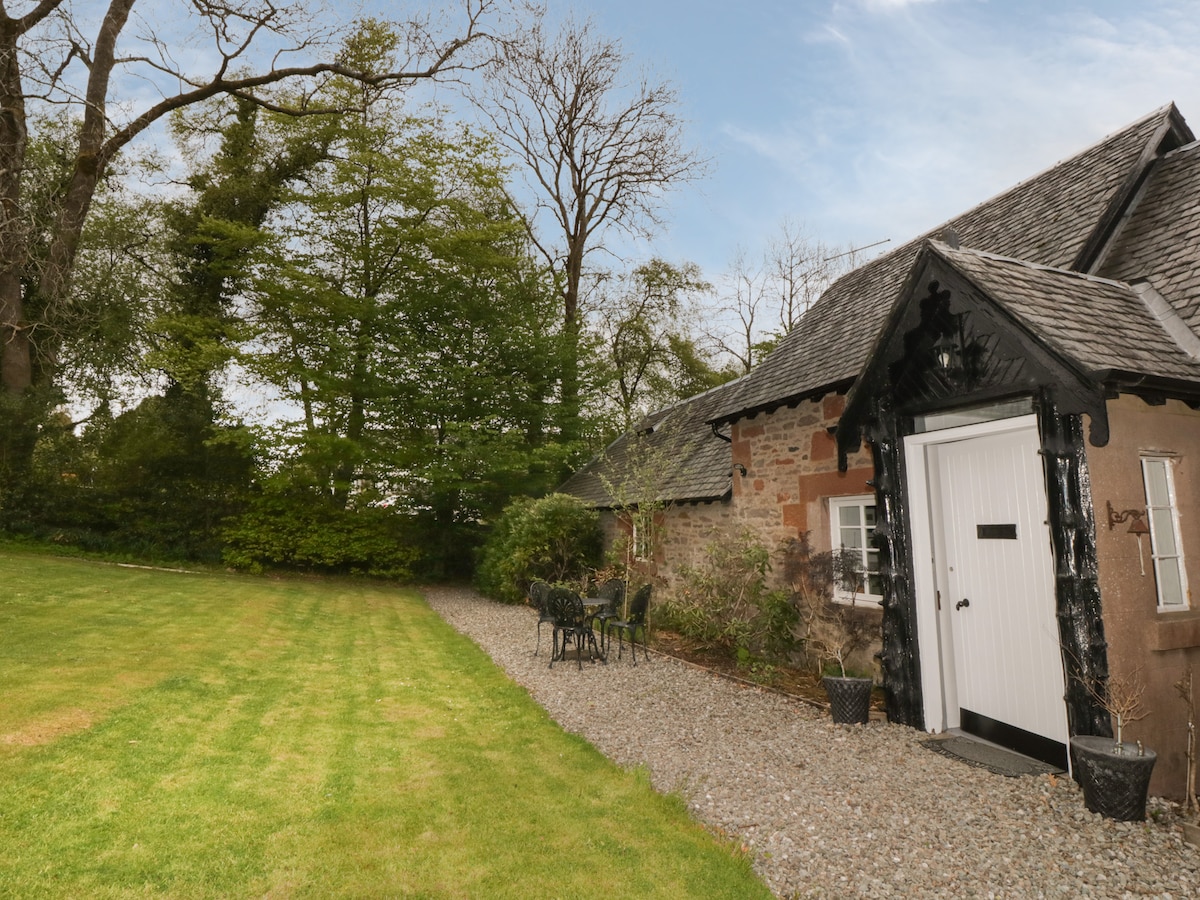 The exterior of the cottage is framed by lush greenery and a manicured lawn. A stone pathway leads to the entrance, flanked by decorative planters. A dining table with several chairs is positioned nearby, providing an inviting outdoor space.