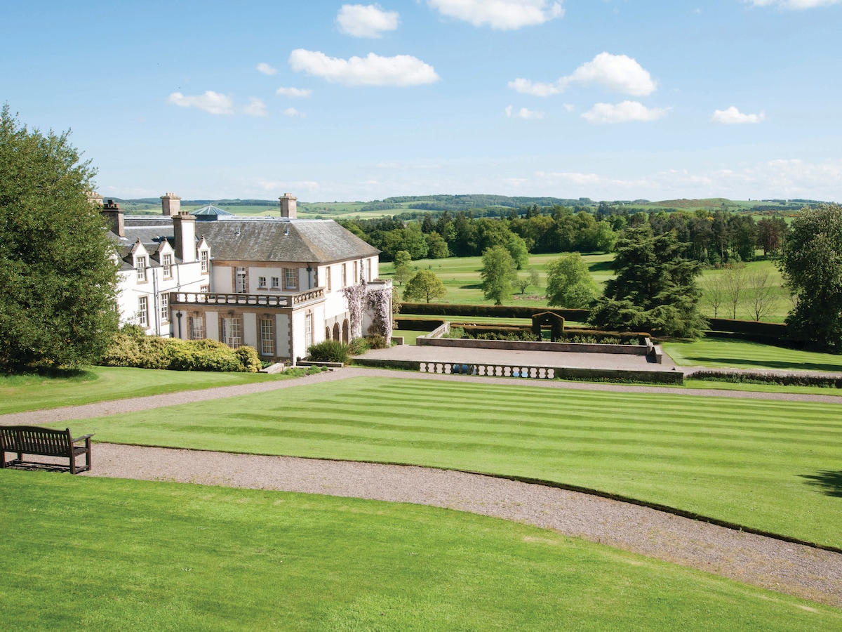 The image captures the elegant exterior of the Edwardian Hill of Tarvit Mansion, set against a backdrop of well-maintained lawns and rolling green hills. A series of neatly arranged terraces lead to a central garden feature, enhancing the tranquil outdoor space.