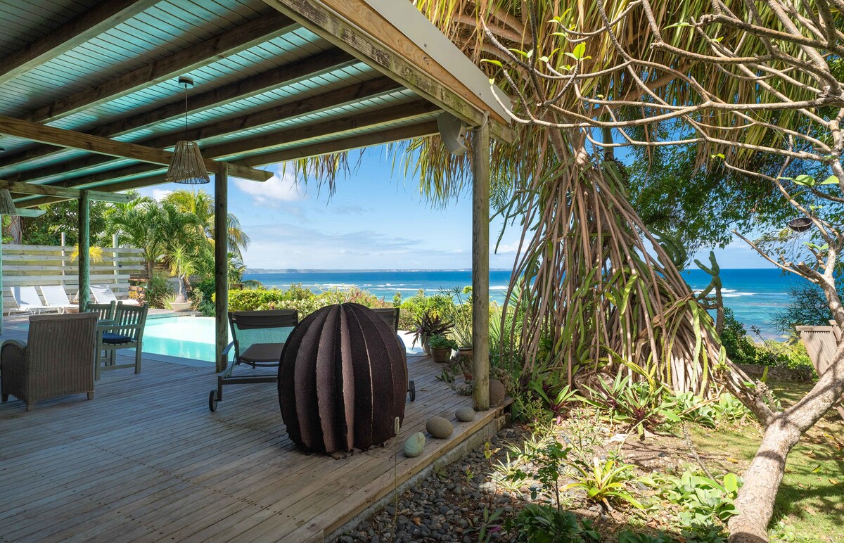 A wooden deck is showcased, surrounded by tropical vegetation and leading to a serene pool. A large round sculpture is visible, complementing the natural landscape. The azure sea stretches in the background under a clear sky, enhancing the peaceful atmosphere.