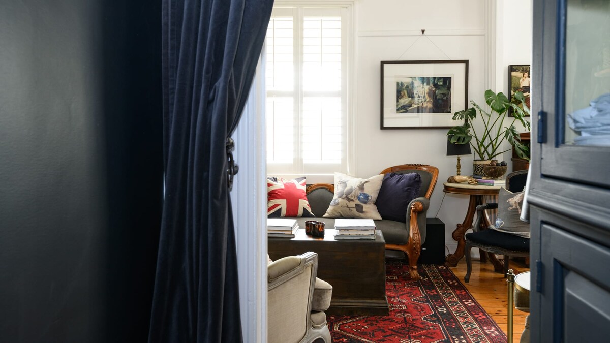 A cozy sitting area is revealed through a doorway, featuring a plush sofa adorned with decorative cushions. A dark wooden coffee table sits on a richly patterned rug, while a potted plant and framed artwork add warmth and character to the space.