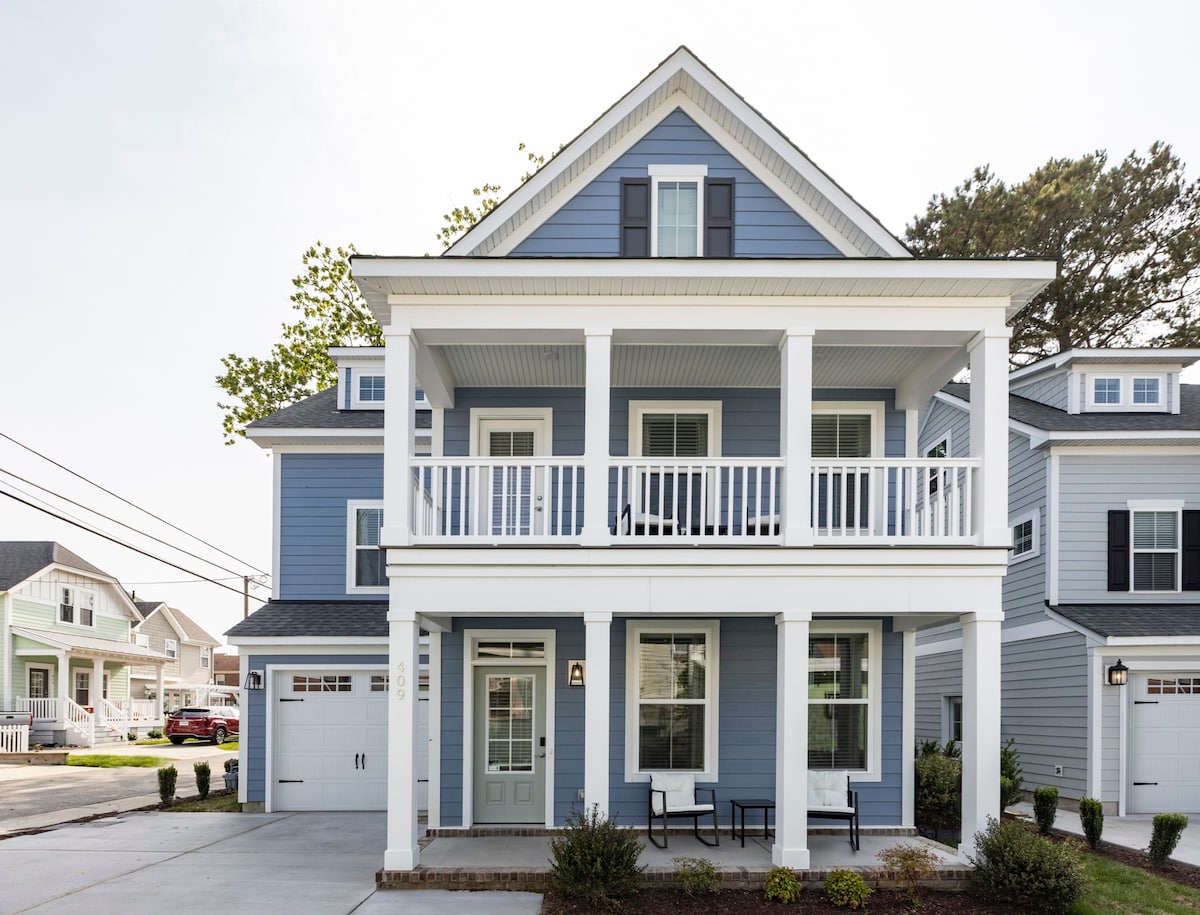 The exterior features a two-story beach house with a light blue facade and white trim. A spacious porch with white railings and dark chairs offers a welcoming entry. Landscaping includes a neatly manicured yard and pathway leading to the front door.
