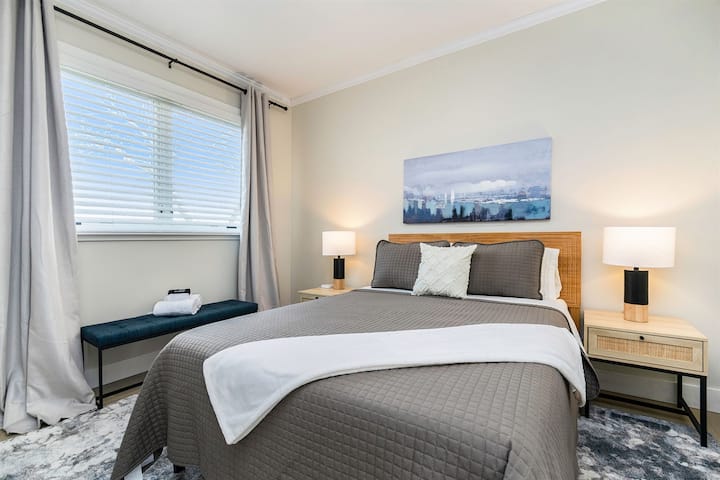 This queen bed room is elegantly designed with black and white bedside lamps, sheer curtains that reach up to the ceiling, and cozy bedding. The room is well-lit with natural light and offers a peaceful ambiance for a restful night's sleep.