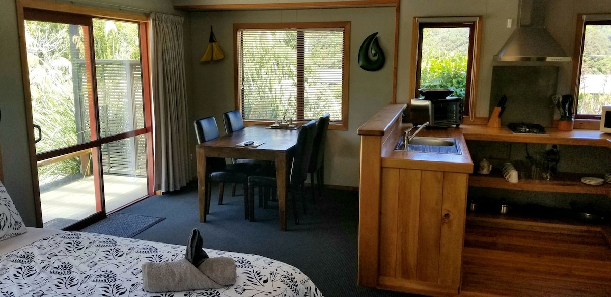 An open living space features a cozy sleeping area with a patterned bedspread, complemented by a table with four chairs. Large windows allow natural light to illuminate the room, showcasing views of the surrounding greenery. A compact kitchen area is visible in the background.