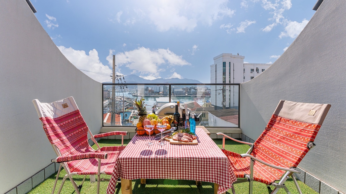 An outdoor terrace is presented with a laid table featuring fruits and beverages. Two folding chairs are positioned on the artificial grass, providing a comfortable spot to enjoy the views of the surroundings and the harbor in the distance, under a bright sky.