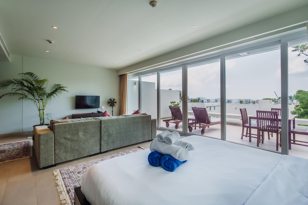 A spacious bedroom is showcased with a large bed featuring rolled towels in blue and white. The living area includes a comfortable couch and a television, while floor-to-ceiling glass doors lead to a private balcony, offering a view of the surroundings.