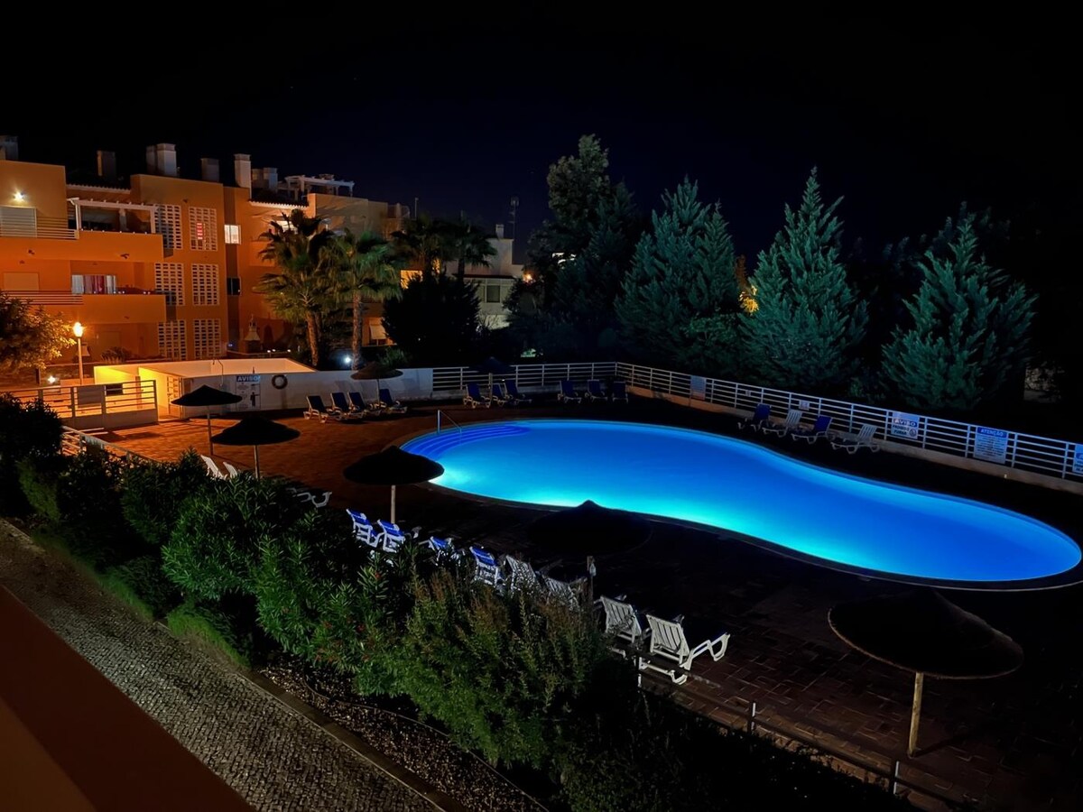 An illuminated swimming pool emits a calming blue glow against a nighttime backdrop. Surrounding the pool, lounge chairs and umbrellas are arranged neatly along the paved area. Lush greenery and trees provide privacy, while soft outdoor lighting contributes to a serene ambiance.