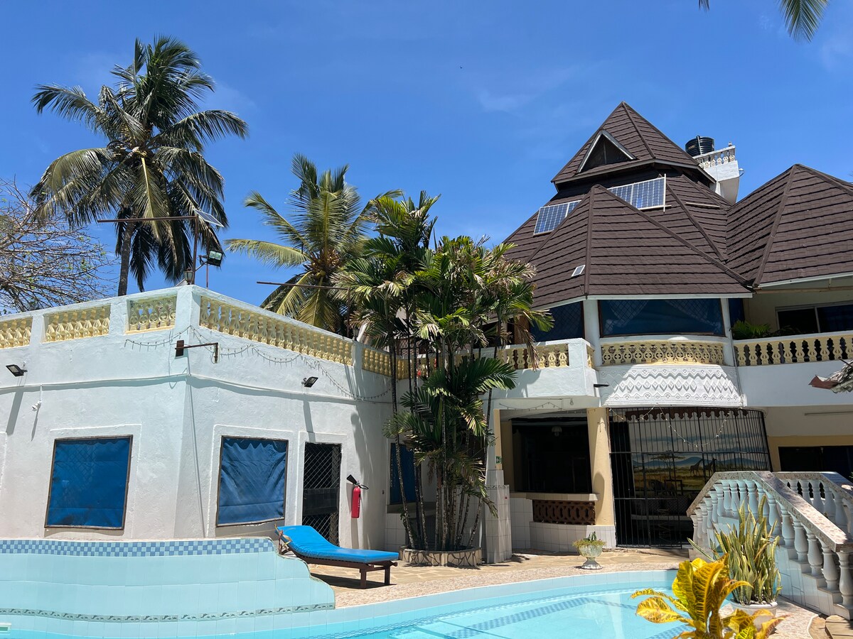 A spacious exterior view of a house surrounded by palm trees and greenery, featuring a multi-level structure with a peaked roof. A bright blue pool is visible in the foreground, with a lounge chair placed beside it. Sunlight brightens the scene under a clear blue sky.