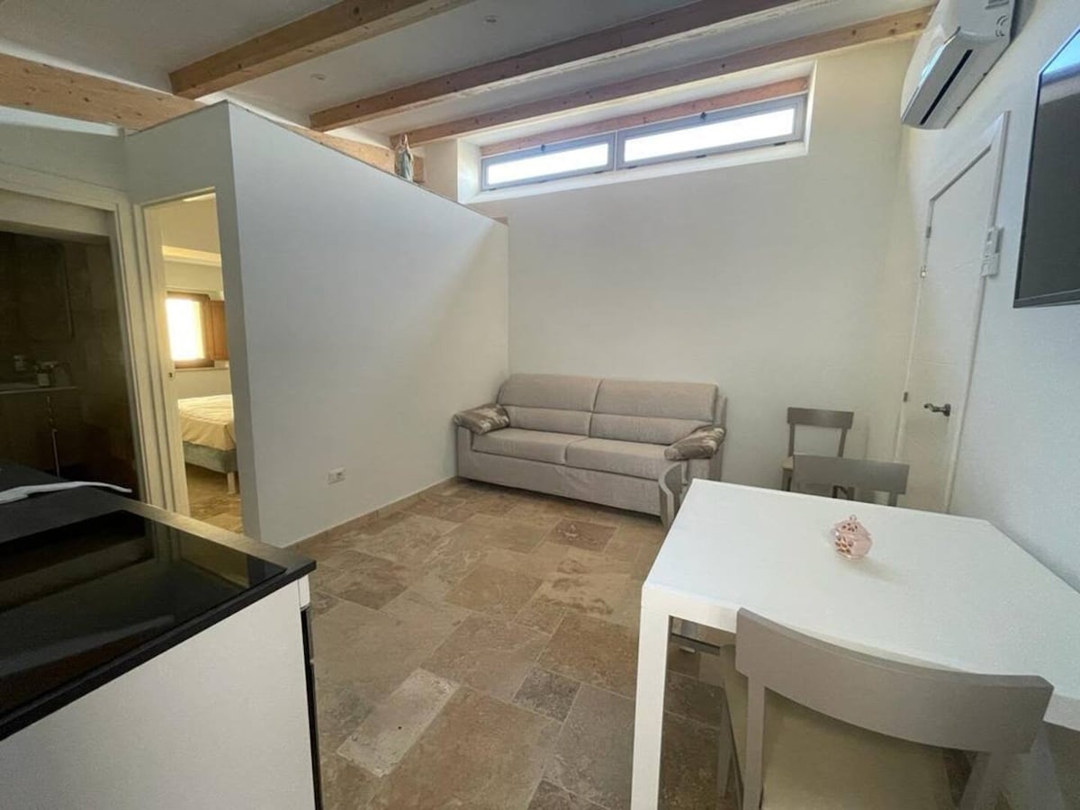 The living area features a light-colored sofa bed accommodating two, paired with a small dining table and chairs. Natural light enters through a set of skylights above, highlighting the stone-tiled floor and the air conditioning unit positioned on the wall.
