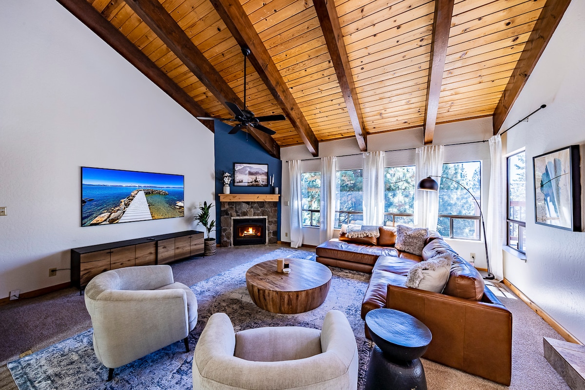 The spacious living room features a vaulted ceiling with wooden beams and large windows offering natural light. A comfortable sectional sofa and accent chairs are arranged around a round coffee table. A wall-mounted TV above a modern fireplace adds to the inviting space.